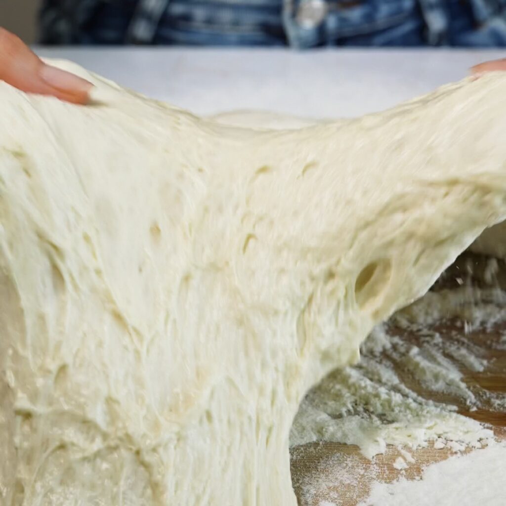 Hands gently neatening chilled bread dough into a square on a generously floured work surface.
