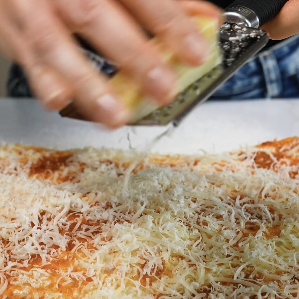 Finely grated Parmesan being sprinkled over sun-dried tomato pesto spread across the bread dough.
