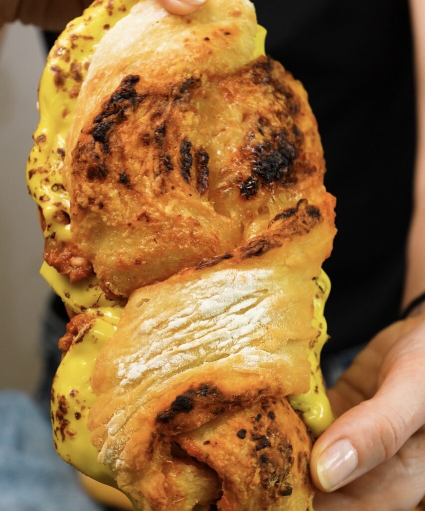 Hands holding a Pesto and Parmesan baguette sandwich filled with beef ragù and melted cheese.