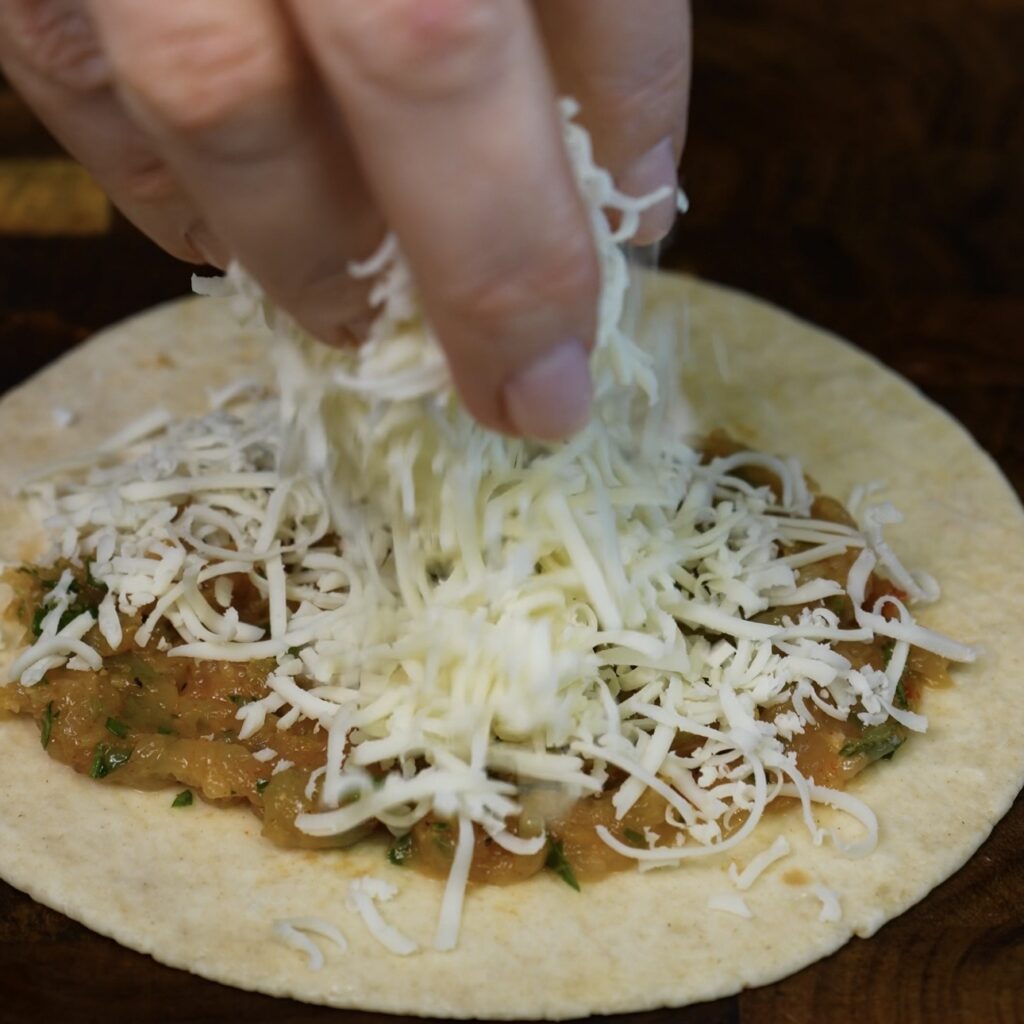 Grated mozzarella being sprinkled over seasoned minced chicken on a mini tortilla.