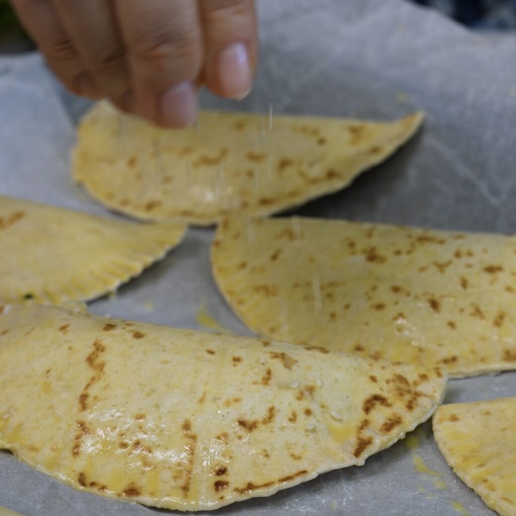 Sesame seeds being sprinkled over glazed tortilla pockets before baking.