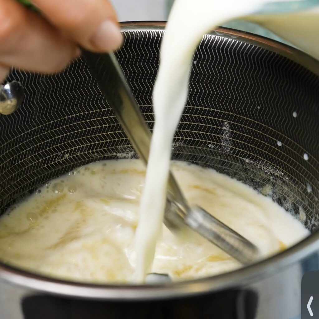 Milk being poured into a saucepan of butter and flour while preparing béchamel sauce.