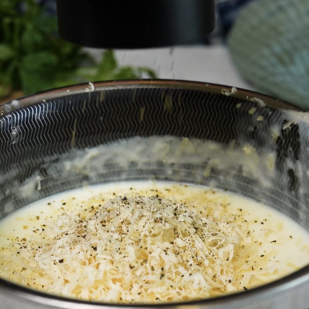 Grated Parmesan being added to a saucepan of creamy béchamel sauce.