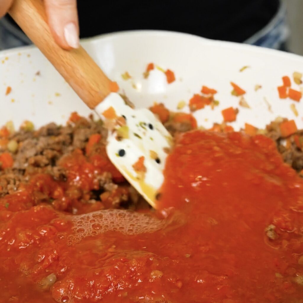 Beef ragù being stirred after chopped tomatoes and water have been added to the pan.