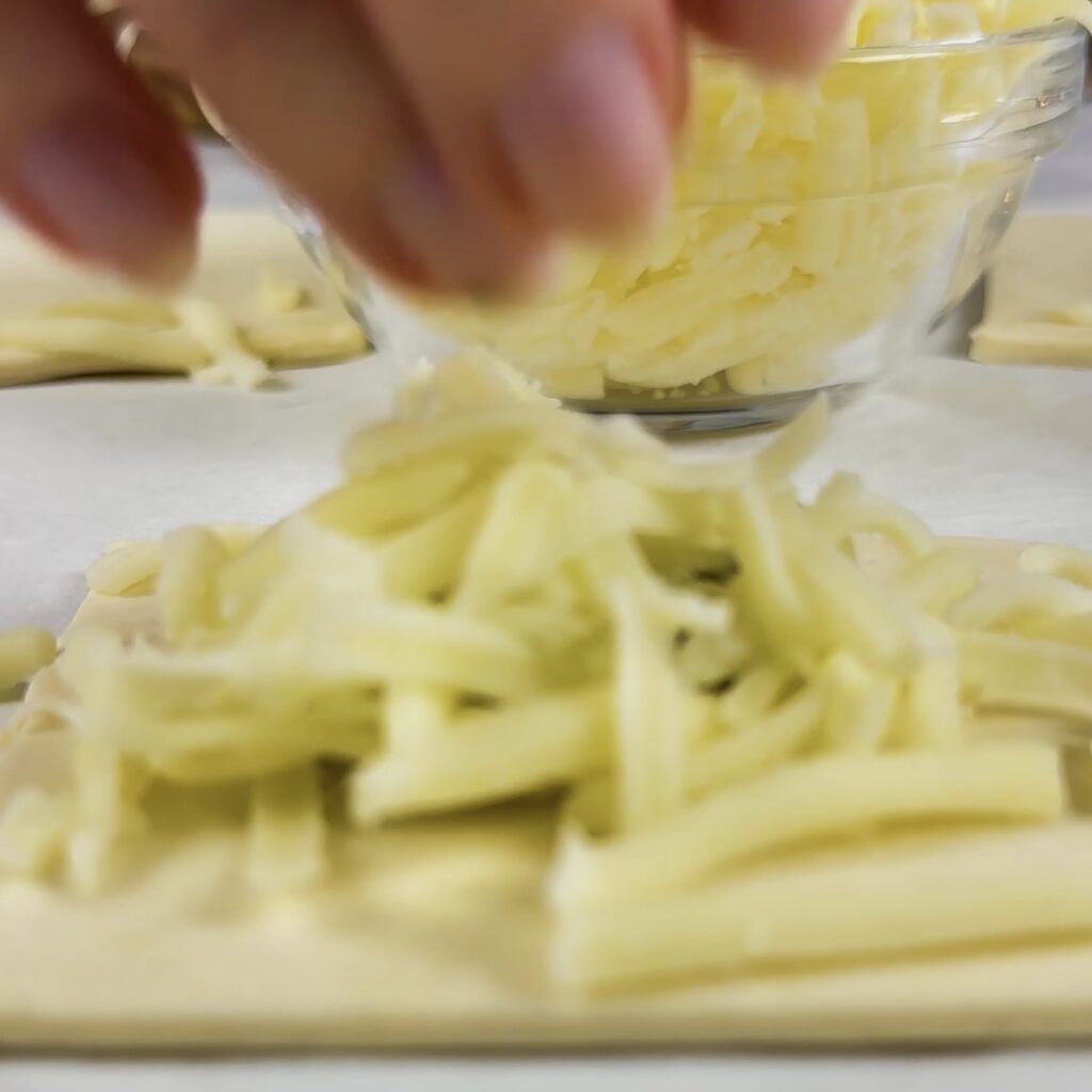 Adding basil pesto and grated mozzarella to puff pastry squares.