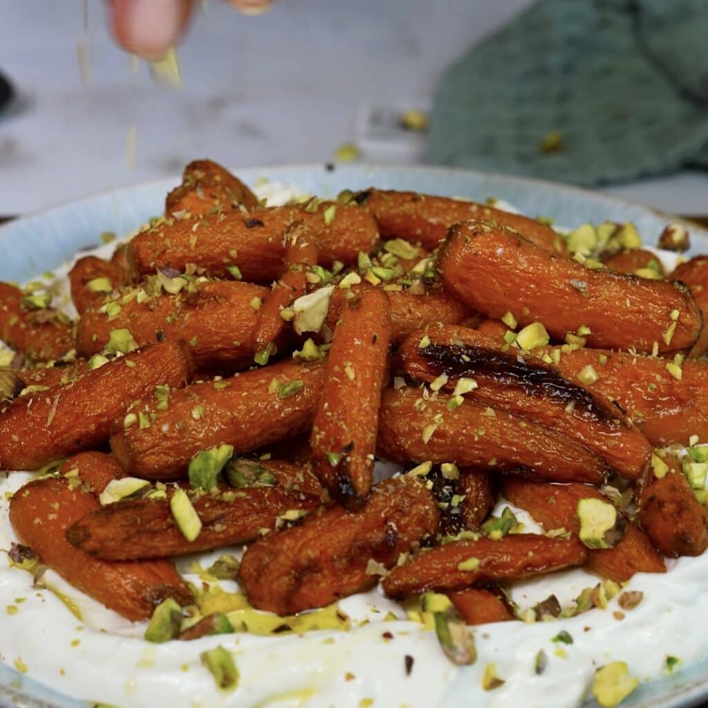 Crushed roasted pistachios being sprinkled over whipped feta and honey-roasted carrots.