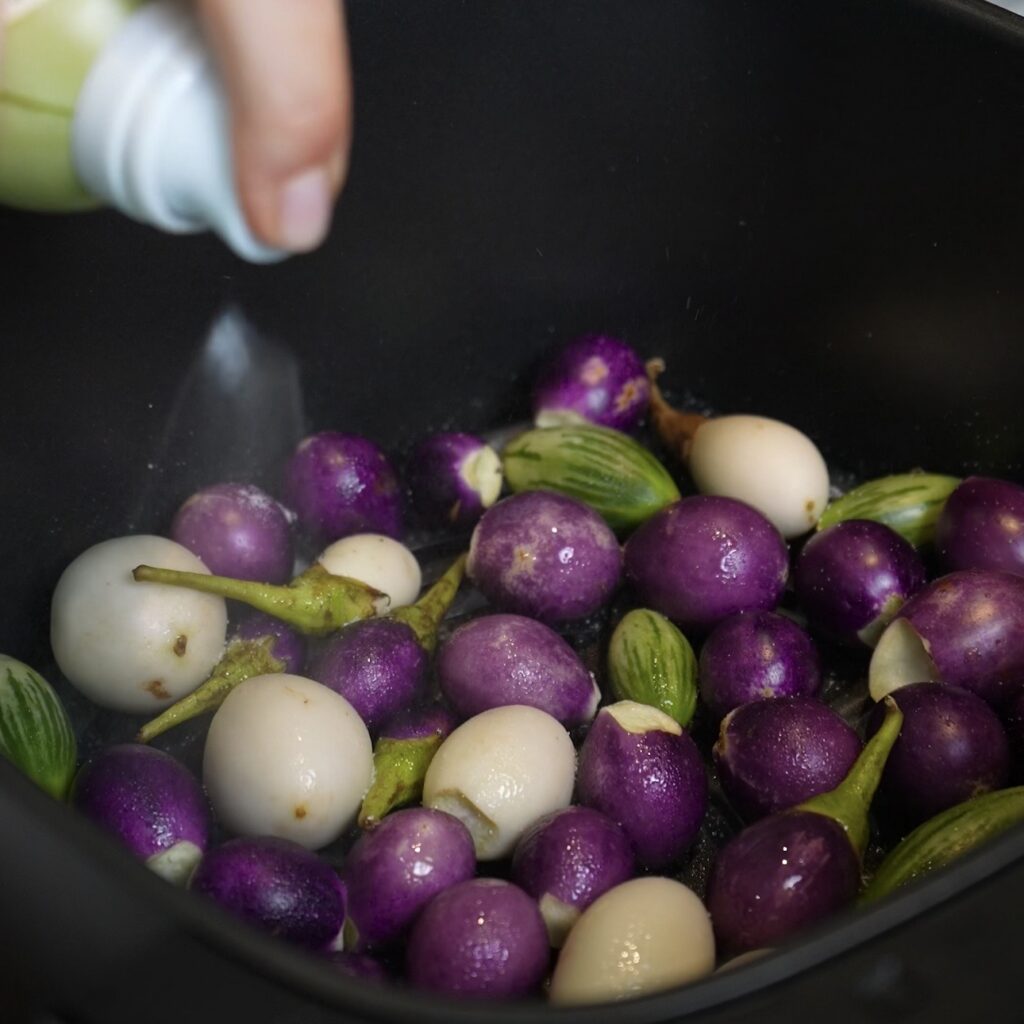 Aubergines in an air fryer basket being sprayed with olive oil before cooking