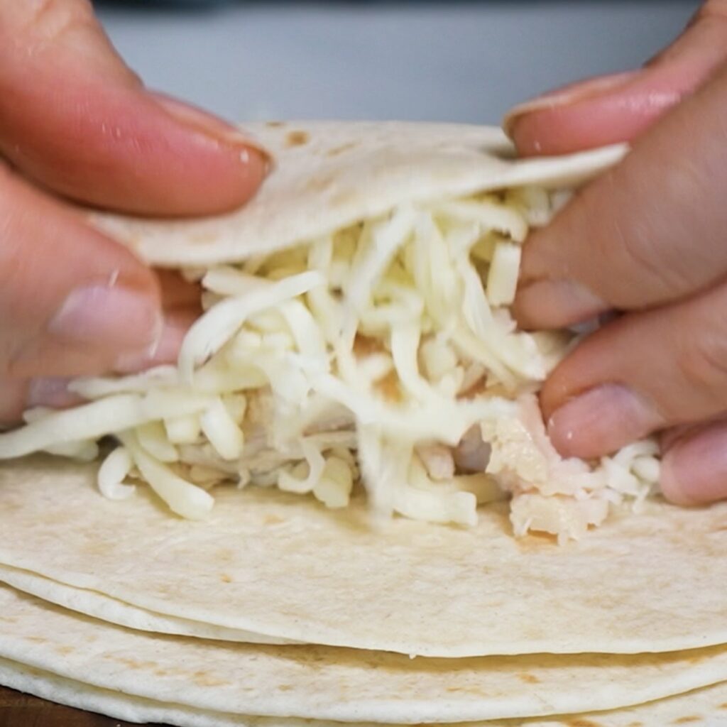 Hands roll a mini tortilla around shredded chicken and grated mozzarella for Cheesy Chicken Tortilla Rolls.