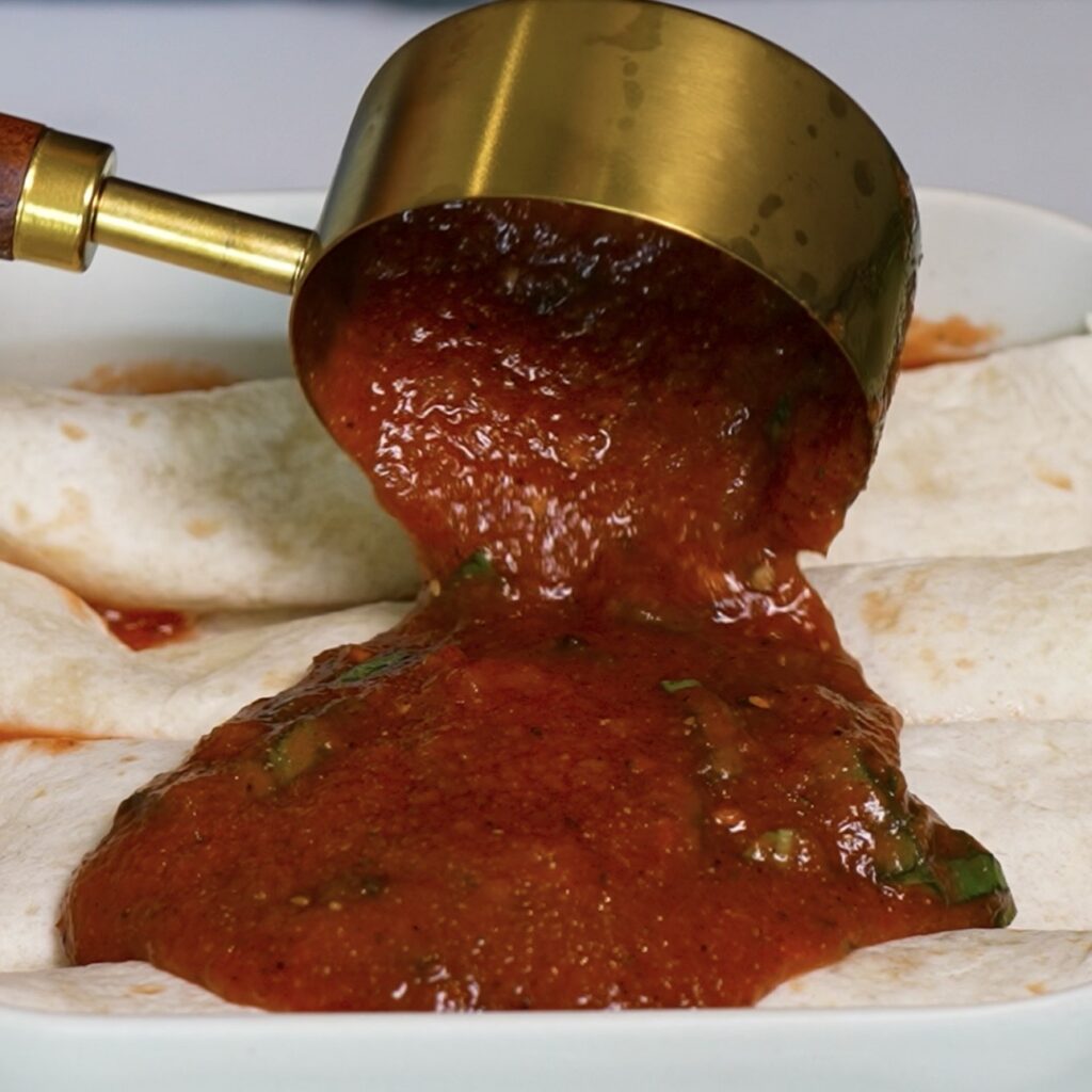 Pouring tomato and basil sauce over chicken tortilla rolls arranged in a square baking dish.