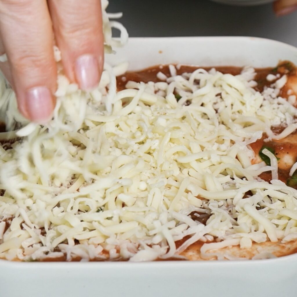 Sprinkling grated mozzarella over chicken tortilla rolls topped with tomato and basil sauce.