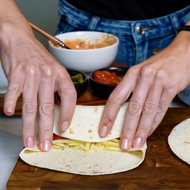 Rolling up a flour tortilla filled with seasoned minced beef, burger sauce, cherry tomatoes, cornichons and grated cheese.