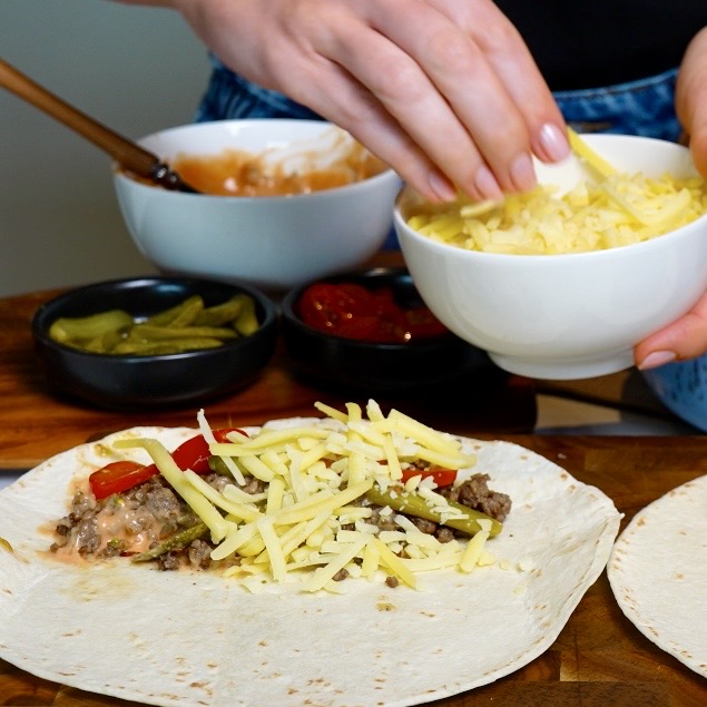 Assembling Cheeseburger Tortilla Rolls with seasoned minced beef, burger sauce, cherry tomatoes, cornichons and grated cheese on a flour tortilla.