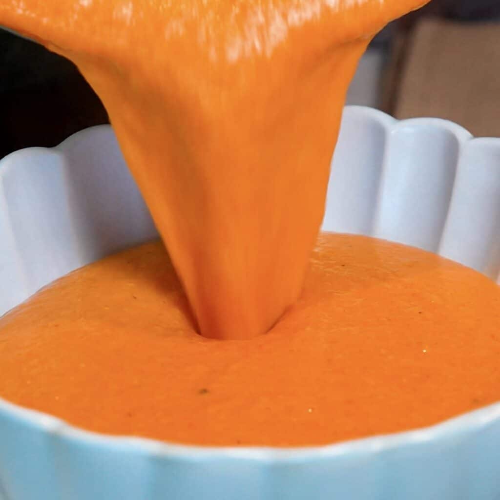 Pouring creamy roasted red pepper and sweet potato soup into a bowl.
