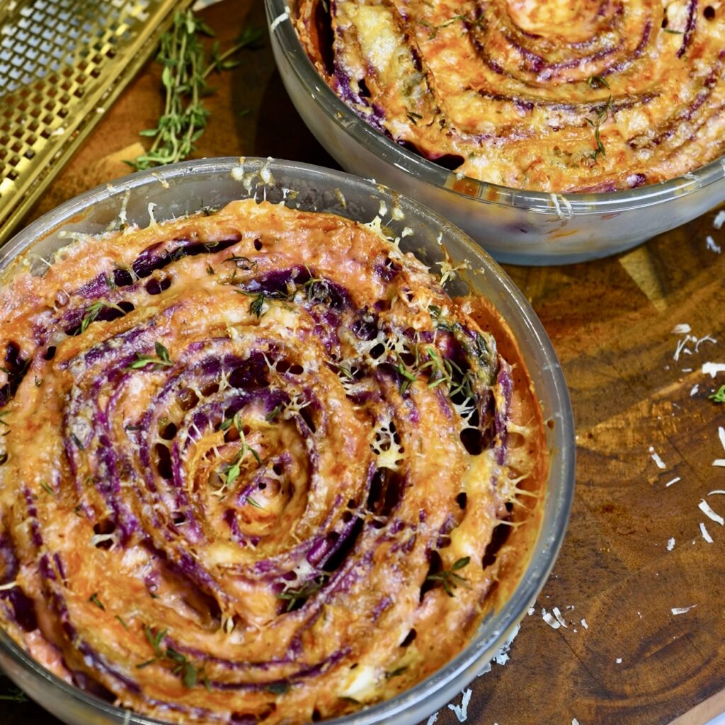 Two finished butternut squash and purple sweet potato gratins in baking dishes on a wooden board.